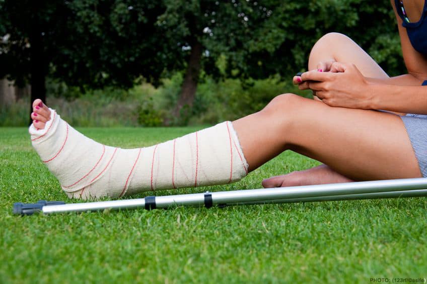 girl with a cast on her broken leg sitting in the grass on her phone. crutches lying down at her side.