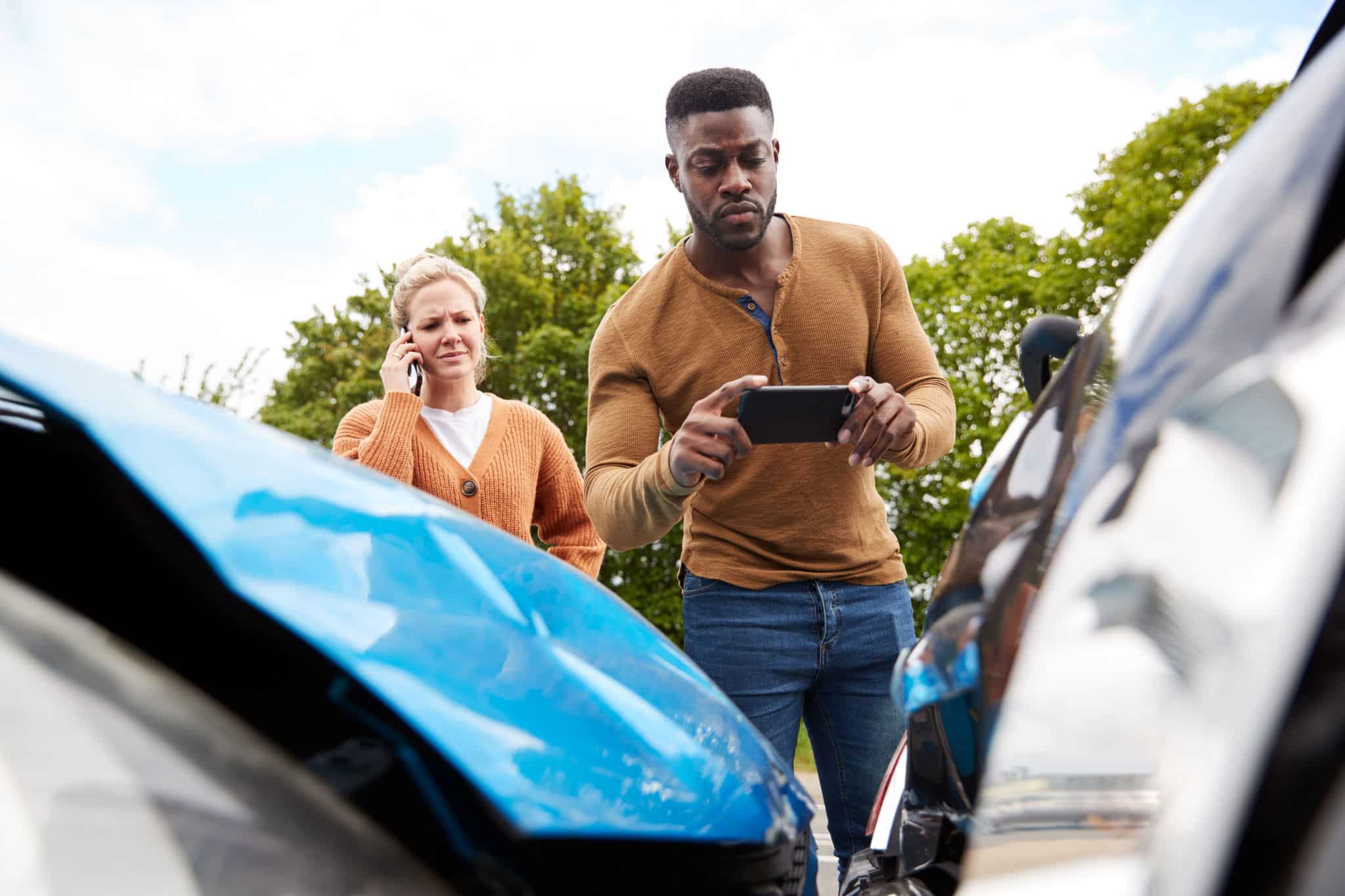 Male driver taking a picture of his car damage after being in a car accident for his insurance claim, with a witness in the background